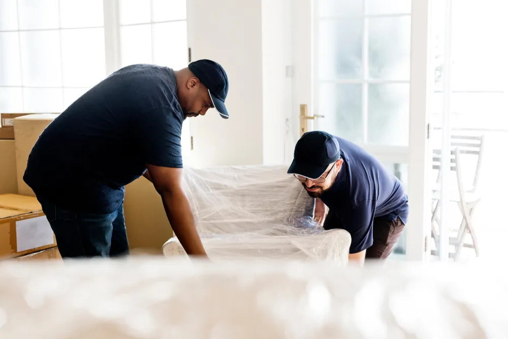 Two movers wrapping furniture in plastic in a bright home